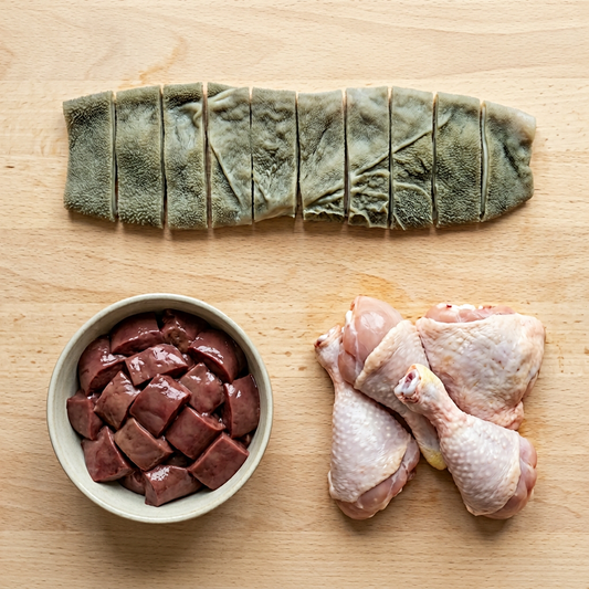 Green tripe, bowl of raw liver, and raw chicken legs on a wooden surface